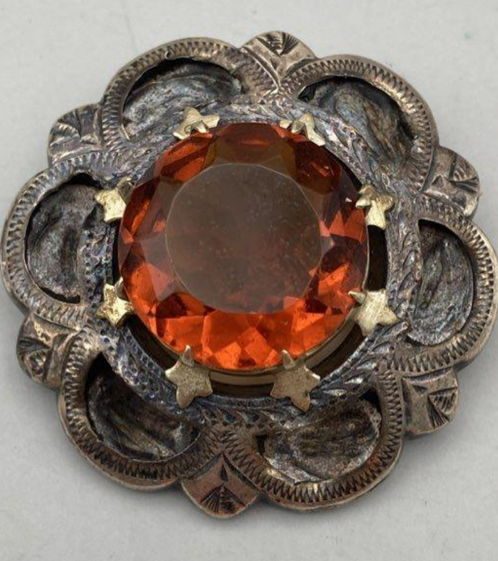 Decorative brooch with a large orange gemstone and intricate metalwork on a light gray background