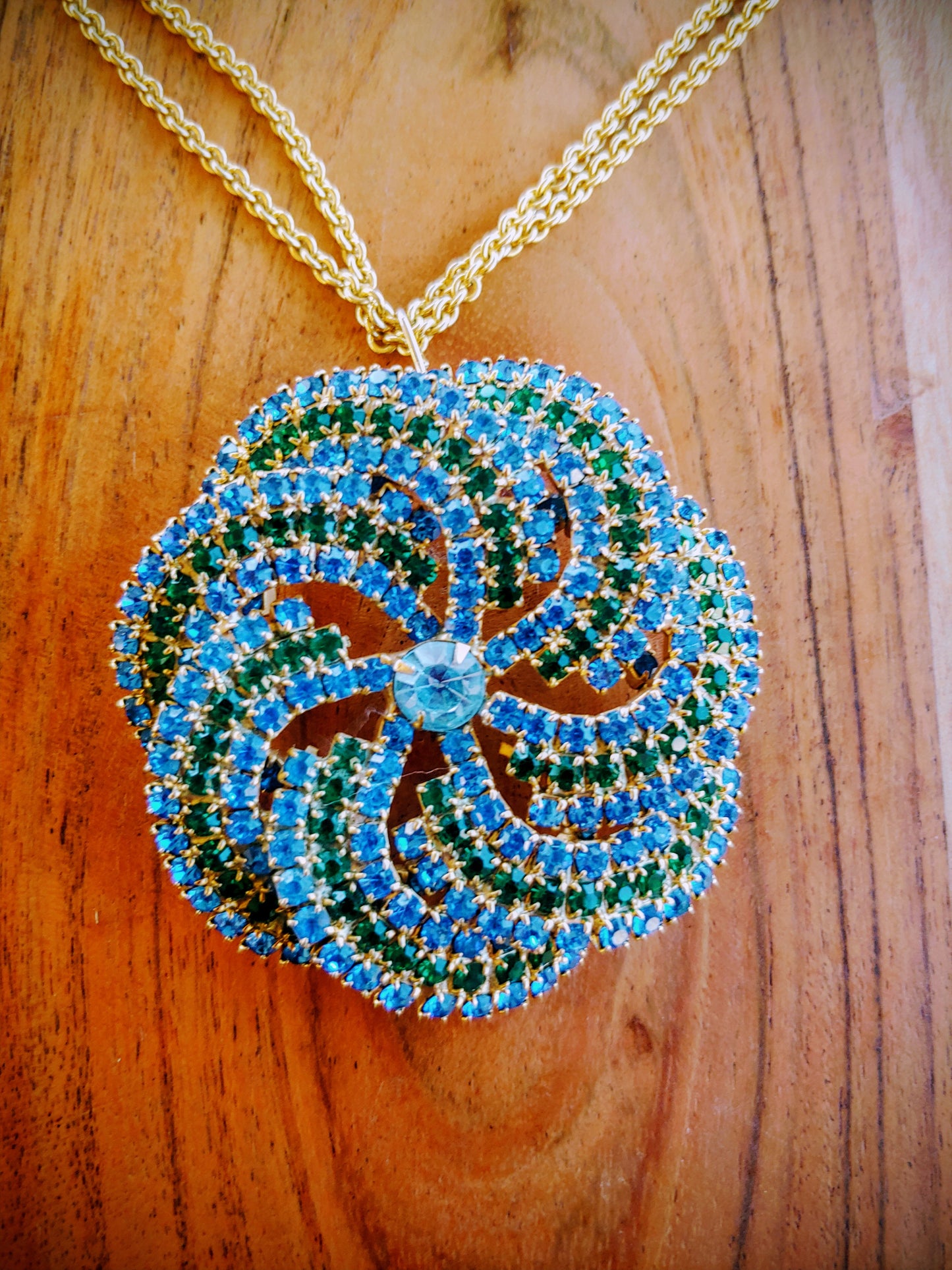Circular pendant with blue and green stones on a gold chain against a wooden background